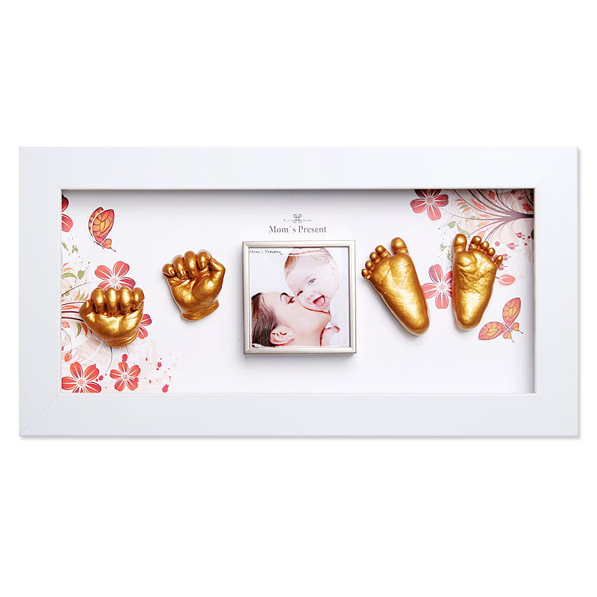 A white rectangular frame with floral designs showcases two golden baby handprints on the left and two golden baby prints on the right. In the center, there is a small photo of a mother kissing a baby, titled "Mom's Present." A lovely Momspresent Baby Hands and Foot 3D Casting Print DIY Kit with White Frame1- The spring of life by LOVE MOM'S PRESENT for cherished memories.