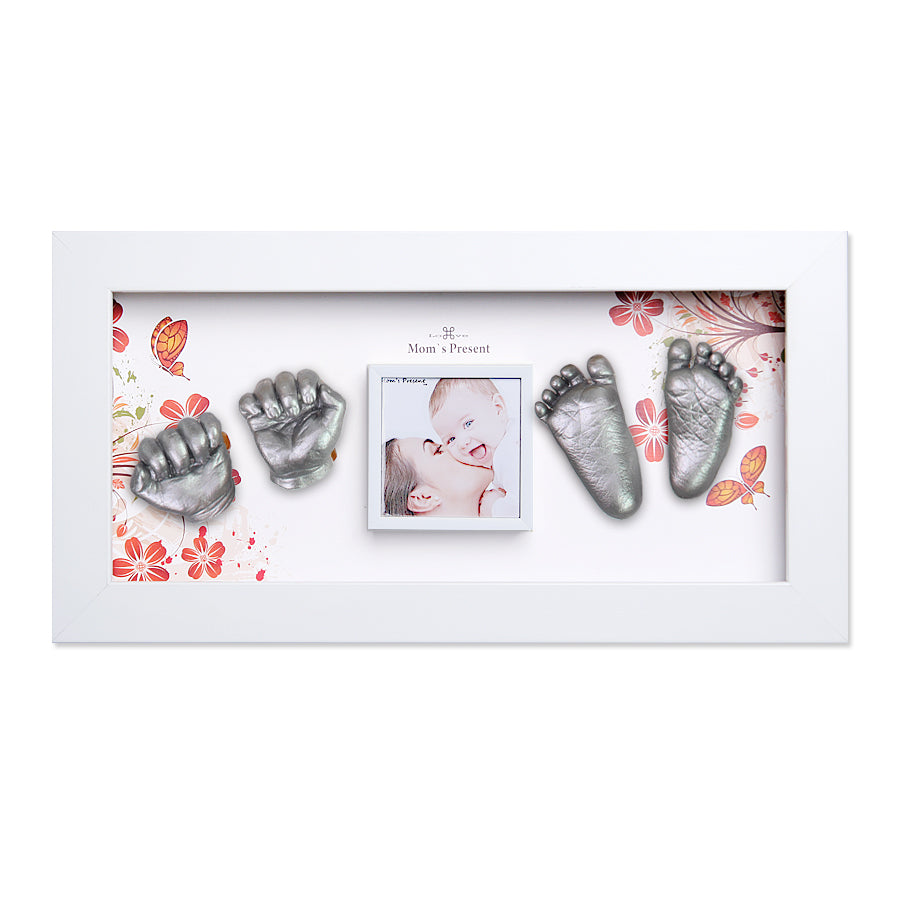 A white rectangular frame displays a baby’s silver hand and foot impressions on a floral background. In the center is a smaller photo of a baby smiling. Text above the photo reads "Mom's Present." This beautiful Momspresent Baby Hands and Foot 3D Casting Print DIY Kit with White Frame1- The spring of life by LOVE MOM'S PRESENT captures precious moments perfectly.