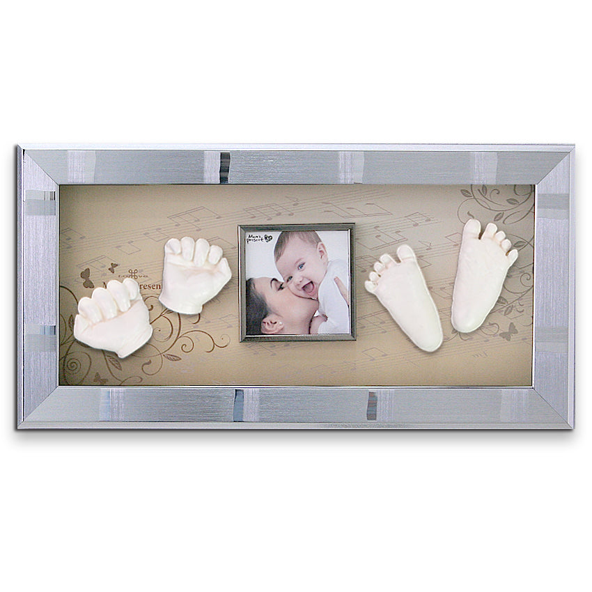 A rectangular shadow box frame displays a photo of a smiling baby and an adult face-to-face. Surrounding the photo are two baby handprints and footprints, all painted white. The frame has a silver border and a background with musical notes and floral designs elegantly completing the charm of the LOVE MOM'S PRESENT Momspresent Baby Hands and Foot 3D Casting Print DIY Kit with SILVER Frame3-piano concerto.