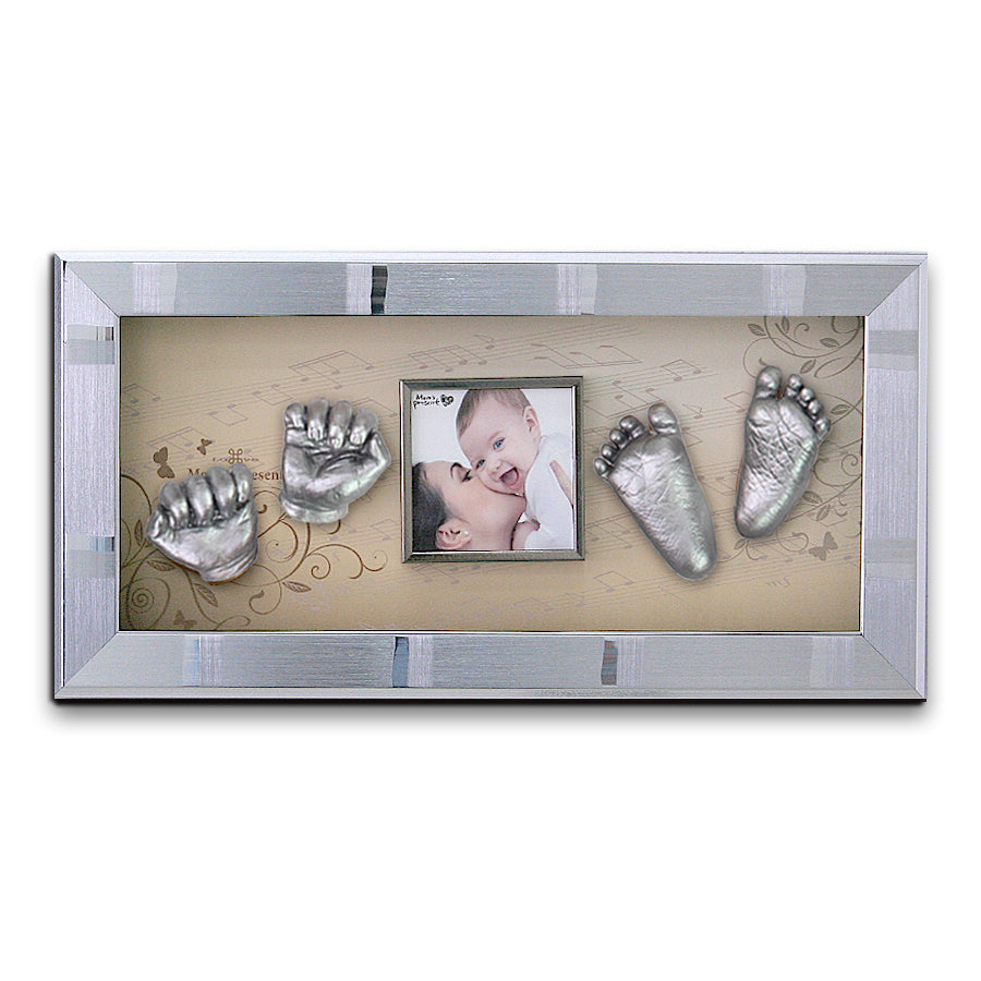 A rectangular frame displaying silver casts of a baby's hands on each side, with a central photo of a smiling baby being kissed on the cheek by an adult. The background features a beige, decorative floral and musical note design. This delightful LOVE MOM'S PRESENT Momspresent Baby Hands and Foot 3D Casting Print DIY Kit with SILVER Frame3-piano concerto makes for a heartwarming memento.