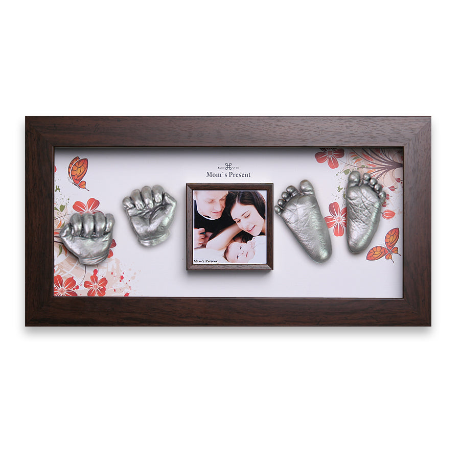 A decorative frame displays a central photo of a mother and baby, surrounded by embossed metallic baby hands and feet. The background features a floral design with the text "Mom's Present" above the photo. Crafted from dark brown wood, this custom baby gift is truly unique and sentimental: the Momspresent Baby Hands and Foot Casting 3D Print DIY Kit with Walnut Frame1 - The spring of life by LOVE MOM'S PRESENT.